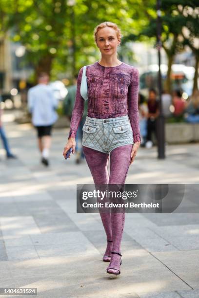 Olga Karput wears gold earrings, a pink and purple denim print pattern long sleeves jumpsuit, blue faded denim quilted shorts, a pale blue matte...