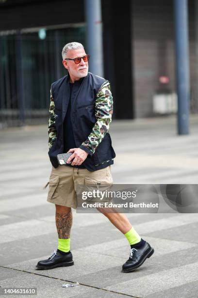 Nick Wooster wears orange sunglasses, a black sleeveless nylon gilet, a khaki / green / brown military print pattern long sleeves nylon t-shirt, a...