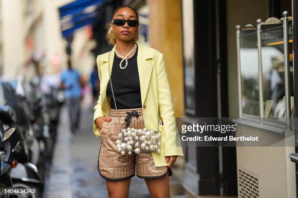 Guest wears black sunglasses, gold snake pendant earrings, white pearls necklaces, a pale yellow long blazer jacket, a black tank-top, a white pearls...