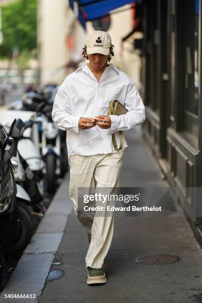 Guest wears a beige with black embroidered pattern Isabelle Marant logo cap, a white shirt, silver rings, a khaki leather zipper clutch, white latte...