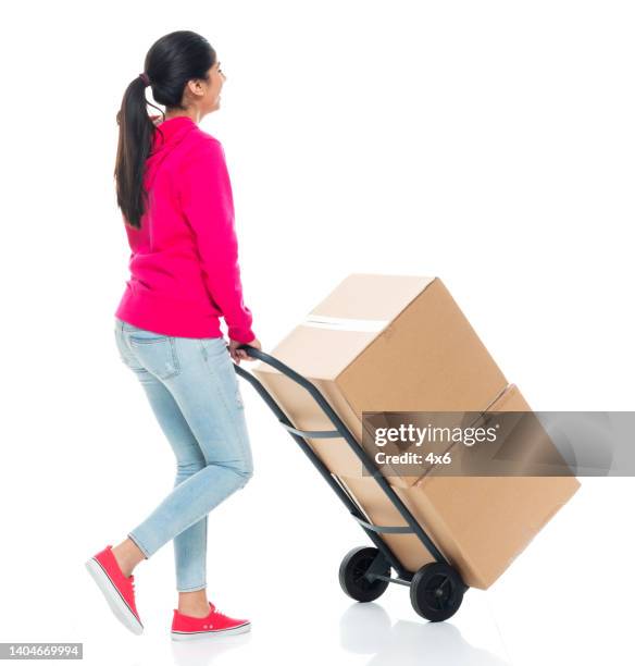 generation z young women manual worker walking in front of white background wearing pants and holding cardboard box and using hand truck - delivery person on white stock pictures, royalty-free photos & images