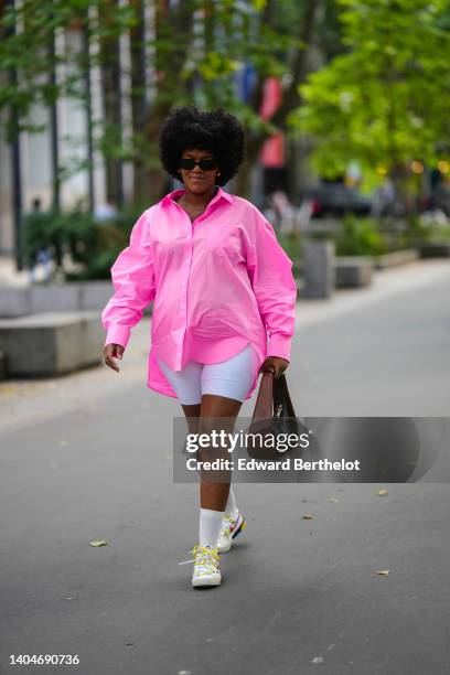 Guest wears black sunglasses, a flashy pink oversized shirt, white cyclist shorts, a khaki and dark brown shiny leather handbag, white socks, white...