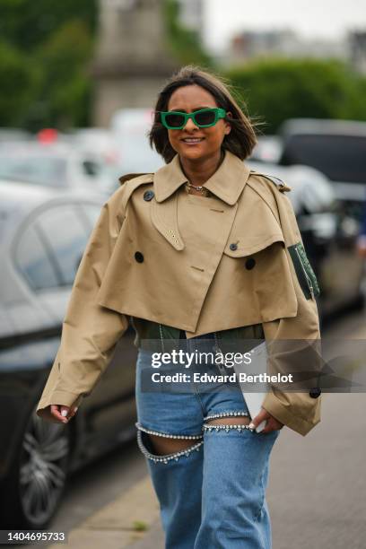 Guest wears green sunglasses from Miu Miu, a silver chain necklace, a beige cropped cloak coat, a khaki shiny leather cropped jacket, blue denim...