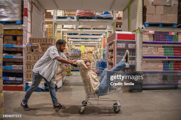 man pushing girlfriend in a trolley down aisle of wholesaler - shopping trolleys stockfoto's en -beelden