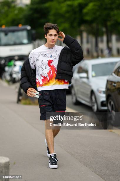 Guest wears a gold chain with blue pendant stone necklace, a white with black embroidered and red / yellow / orange fire print pattern pullover from...