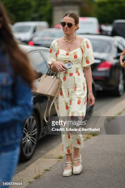 Guest wears black sunglasses in shape of heart, gold necklace, a pale yellow with red pineapple print pattern V-neck / puffy sleeves jumpsuit, a...