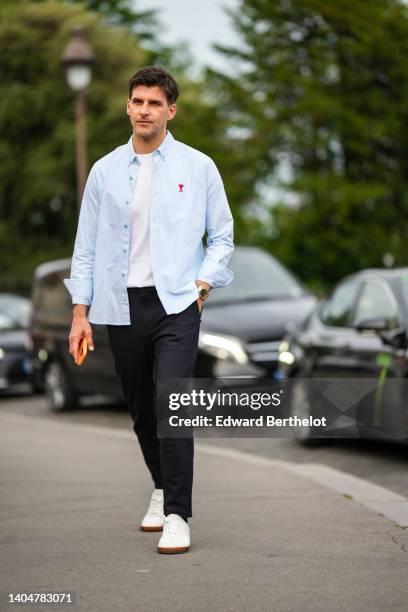Johannes Huebl wears a white t-shirt, a pale blue open shirt with embroidered red logo shirt from Ami, black sunglasses, white leather sneakers, a...