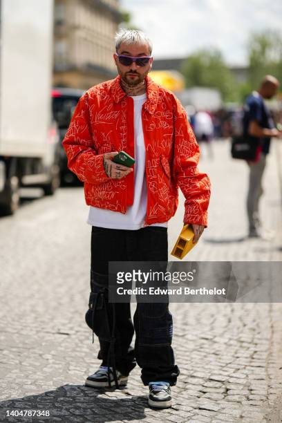 Maxime Chastan wears purple sunglasses, silver earrings, a white t-shirt, a red with embroidered white oversized jacket, black denim cargo pants, a...