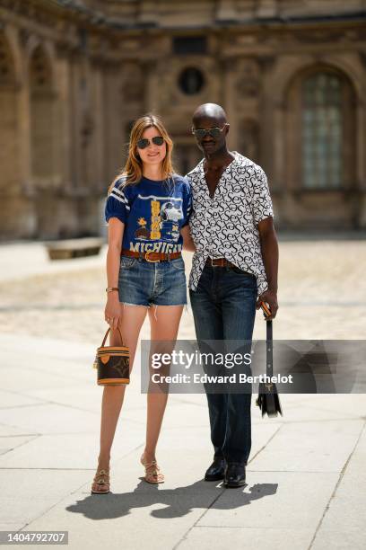 Eugénie Trochu wears black sunglasses from Ray Ban, silver earrings, a navy blue with white and yellow print pattern t-shirt, a camel shiny leather...