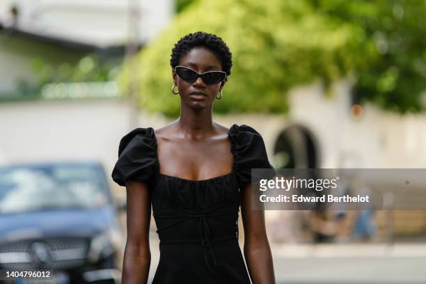 Guest wears black sunglasses, gold earrings, a black square-neck / puffy short sleeves / belted short dress, outside the Amiri show, during Paris...
