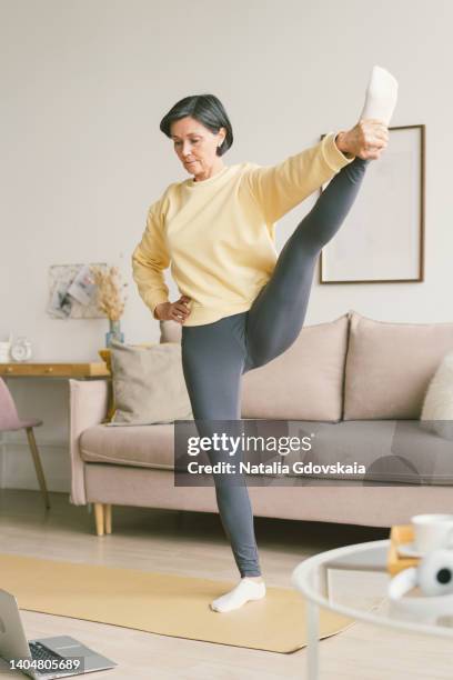 elderly old lady stretches hands with feet holding leg. balancing yoga exercise, wellness for retired female. senior woman stretching, online training with laptop in living room. exercising for health. well-being, wellness for retired female. home fit - camel active fotografías e imágenes de stock