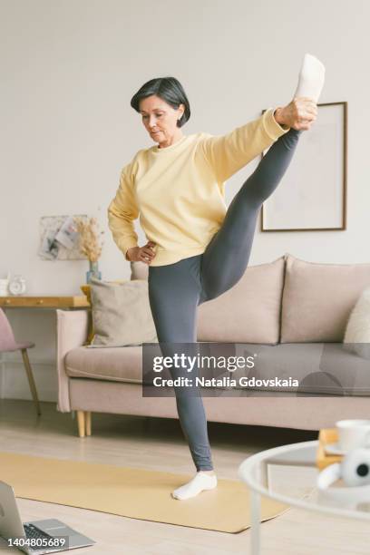 elderly old lady stretches hands with feet holding leg. balancing yoga exercise, wellness for retired female. senior woman stretching, online training with laptop in living room. exercising for health. well-being, wellness for retired female. home fit - camel active stockfoto's en -beelden