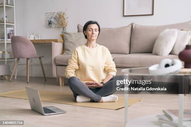 retired woman meditating and practicing yoga while sitting in lotus pose on floor at home. peaceful senior woman in lotus position meditation with closed eyes at home while sitting on yoga mat on floor. calm elderly lady practicing home workout - senior inhaling stock pictures, royalty-free photos & images