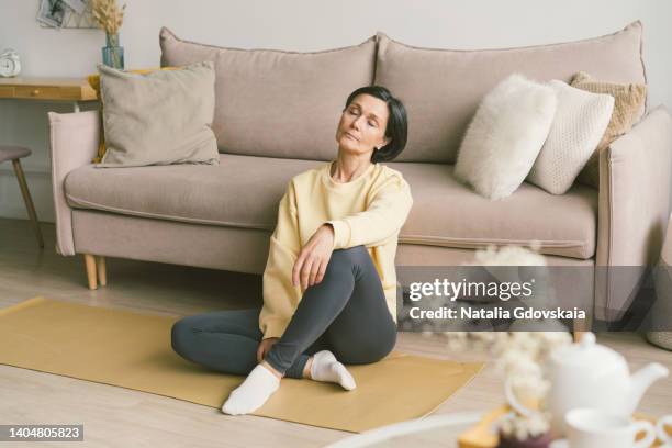 senior fatigue woman with closed eyes sitting on yoga sport mat in living room. relax after exercising fitness. emotional and spiritual health. well-being, wellness with meditation. enjoying good moments, slow life and niksen - camel active - fotografias e filmes do acervo