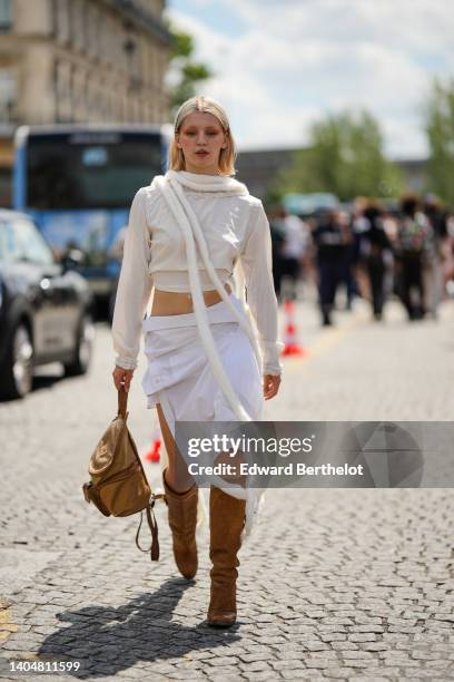 Guest wears a silver piercing, a white fluffy long scarf, a white latte ruffled long sleeves cropped t-shirt, high waist white wrap knees skirt, a...