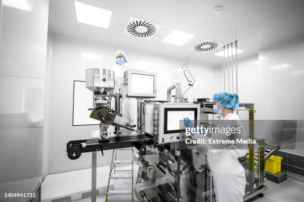 frau in einem labor mit schutzhandschuhen, maske, mütze und anzug vor dem bildschirm der maschine in einer pharmazeutischen fabrik gesehen - arzneimittelherstellung stock-fotos und bilder