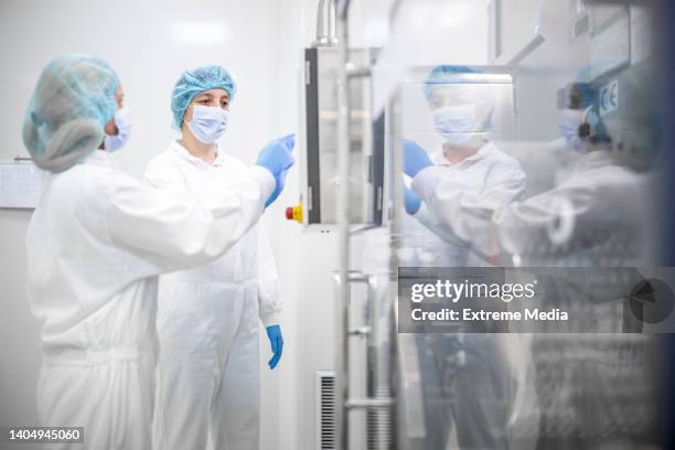 female employees in a pharmaceutical laboratory wearing proper equipment seen while pushing a button on the machine in a laboratory during pharmaceutical manufacturing - cleanroom stock pictures, royalty-free photos & images