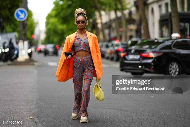 Debbie @blackhey wears black sunglasses, an orange oversized blazer jacket, a blue / green / pink / brown print pattern cropped shirt, gold earrings,...