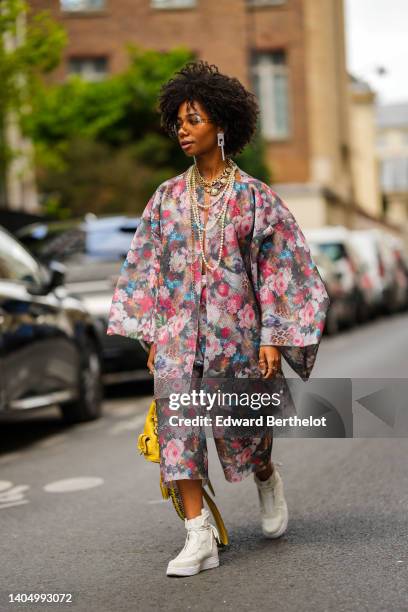 Sarah Monteil wears sunglasses, large plastic pendant earrings, large gold chain necklace, white pearls necklace, a multicolored flower print patter...