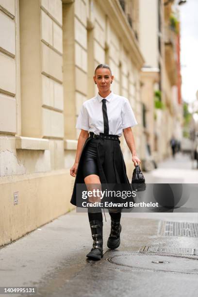 Alice Barbier wears a gold and black earring, diamond earrings, a white short sleeves shirt, a black silk tie, a black denim slit / belted / pleated...