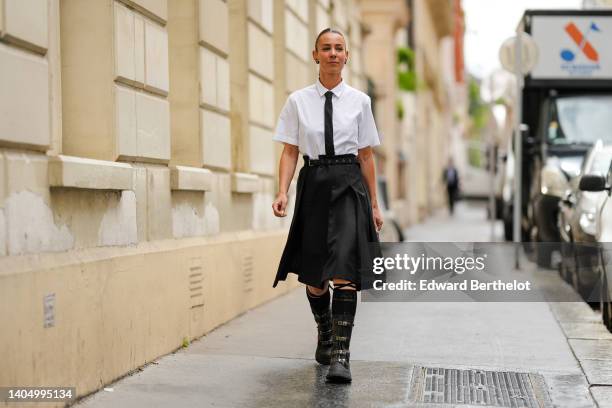 Alice Barbier wears a gold and black earring, diamond earrings, a white short sleeves shirt, a black silk tie, a black denim slit / belted / pleated...