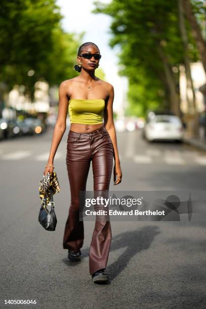 Guest wears black sunglasses, gold large earrings, a gold chain pendant necklace, khaki shiny satin shoulder-off cropped top, brown shiny leather...