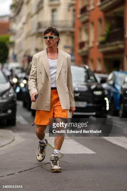 Guest wears green sunglasses, a beige long blazer jacket, a white tank-top, orange shorts, white socks from Nike, beige and burgundy print pattern...