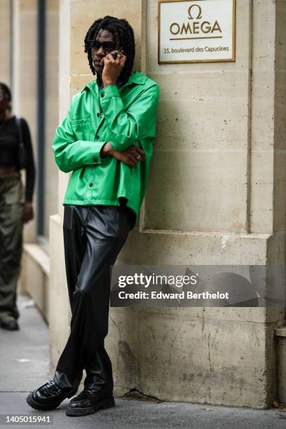 Guest wears black sunglasses, silver rings, a black t-shirt, a silver chain necklaces, a green shiny leather jacket, black shiny leather large pants,...