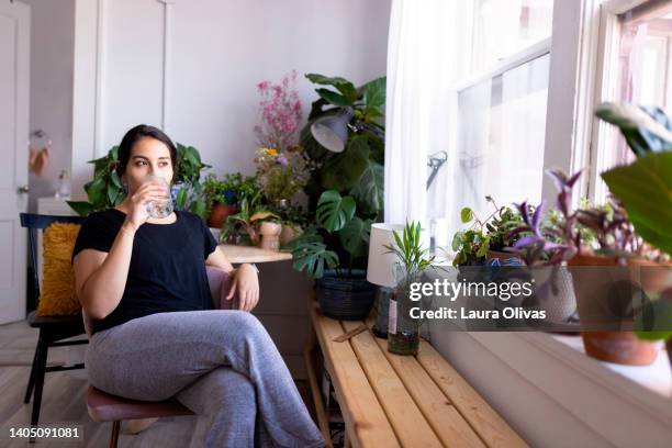 young adult woman sitting by a window drinking from a glass of water - drinking water stock pictures, royalty-free photos & images