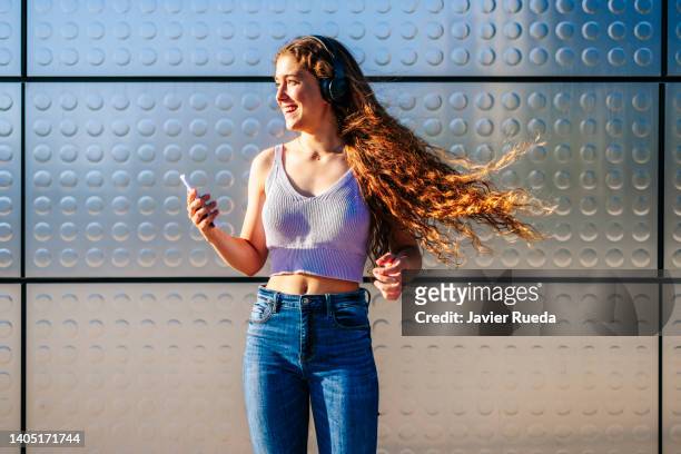 candid portrait of authentic positive teen listening happy music and dancing. gorgeous young girl with headphones and long curly ginger hair having a good time outdoors. adolescence street lifestyle concept. copy space. - mens long jump stockfoto's en -beelden