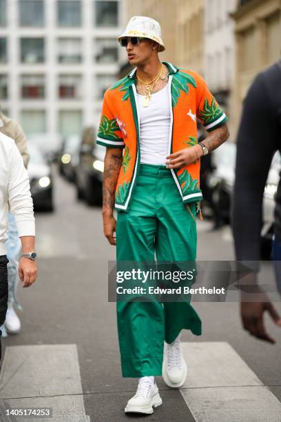 Kyle Kuzma wears a white with yellow and blue print pattern bob, sunglasses, gold long chain necklaces, a white ribbed tank-top, an orange with green...