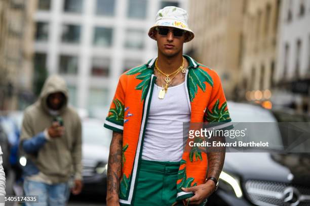 Kyle Kuzma wears a white with yellow and blue print pattern bob, sunglasses, gold long chain necklaces, a white ribbed tank-top, an orange with green...