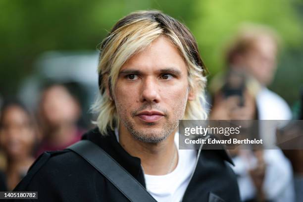 Orelsan wears a white t-shirt, a silver chain necklace, a black zipper jacket, a black shiny leather crossbody bag , outside the Loewe show, during...