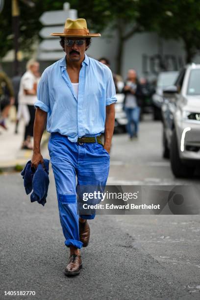 Haider Ackermann wears a pale yellow and brown suede / felt hat, a white tank-top, a baby blue shirt, a khaki braided suede belt, royal blue large...