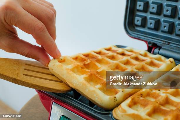a woman or a girl takes a freshly cooked and baked waffle in a waffle iron to eat it. the concept of cooking homemade food according to a traditional family recipe, hobby, favorite activity. dependence on eating sweets and desserts, overeating. - waffles stock pictures, royalty-free photos & images