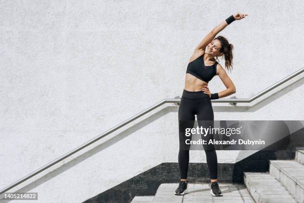 happy young woman runner in sportswear doing warm-up before morning run smiling enjoying workout against white wall outdoor in city street. copy space. - workout gear stock pictures, royalty-free photos & images