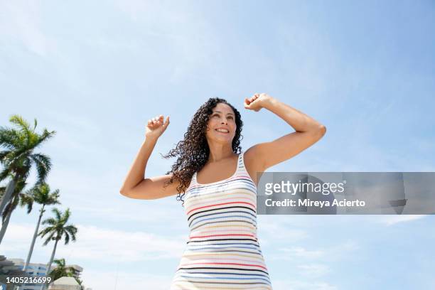 a latinx woman with a long curly hair, happily dances with her arms up in the air in front of a blue sky, while wearing a striped dress. - mature latin women fotografías e imágenes de stock