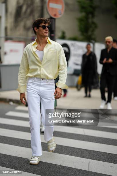 Guest wears black sunglasses, a white tank-top, a pale yellow shirt, white denim jeans pants, white socks, beige strappy velcro sandals, outside the...