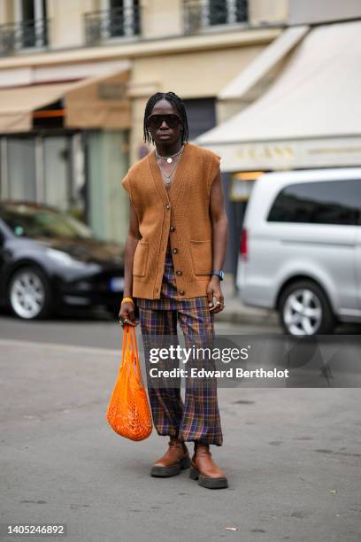 Guest wears black sunglasses, silver chain pendant necklace, a brown sleeveless oversized cardigan, a navy blue Apple watch, brown and navy blue...