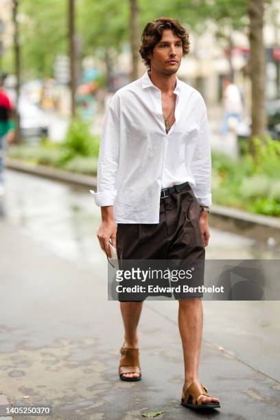 Guest wears a white V-neck shirt, a silver long chain pendant, a black shiny leather belt, dark brown suit pants, brown shiny leather sandals,...