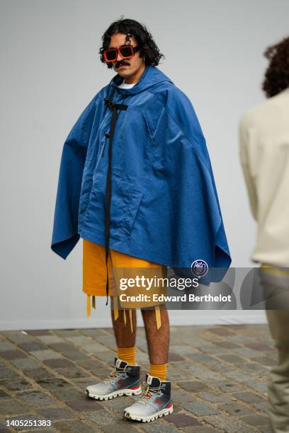 Guest wears orange sunglasses, a blue denim hoodie oversized cloak, yellow orange fringed shorts, yellow socks, white and black gradient print...