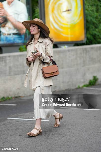 Guest wears a beige wicker hat, pearl earrings, a beige short trench coat, a brown shiny leather handbag from Ralph Lauren, a beige midi dress, brown...