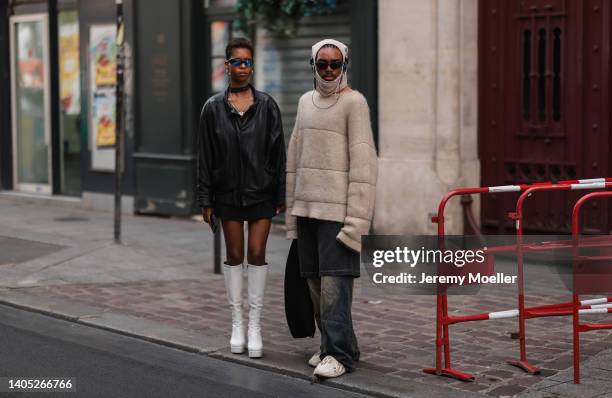 Guest wears blue sunglasses, a silver necklace, silver earrings, a black leather bomber jacket, a black mini skirt and white shiny leather high...