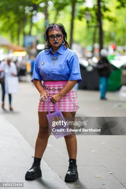 Guest wears black glasses, silver earrings, a blue short sleeves shirt from YSL / Saint Laurent, a gold and white brooch, a silver pendants necklace,...