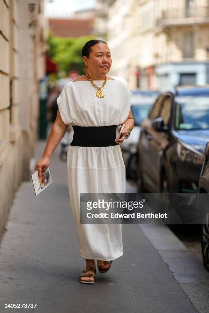 Guest wears a gold large chain pendant necklace, a white short sleeves / large belt / long dress, gold bracelets, a silver watch, gold rings, gold...