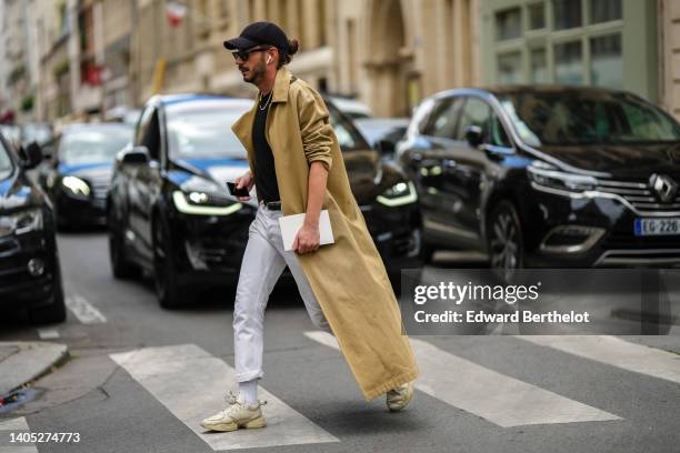 Guest wears a black cap, black sunglasses, a black t-shirt, a silver chain necklace, a beige long trench coat, white denim large jeans pants, a black...