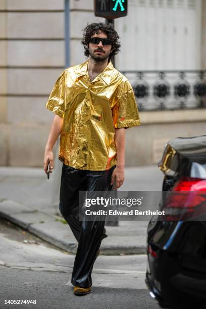 Guest wears black sunglasses, a gold shiny varnished leather short sleeves shirt, black shiny leather large pants, brown suede pointed ankle boots ,...