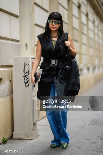 Guest wears silver sunglasses, a silver chain necklace, a black sleeveless oversized long gilet, a black belt, a black jacket, blue faded denim large...