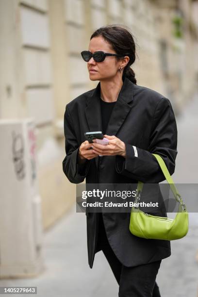 Guest wears black sunglasses, a black t-shirt, a black oversized blazer jacket, a green shiny leather handbag, black denim jeans pants , outside the...