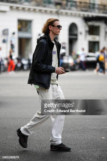 Guest wears black sunglasses from Versace, a beige with embroidered navy blue pattern sweater, a black hoodie rain coat, white gray denim jeans...